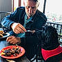 man, dog, food, fork, plate, table, denim_jacket, cap, red_harness, indoor, window, chair, meal, black_dog, person, pet, animal, waiting, casual, restaurant