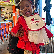 Sarai joined the competition — help win amazing prizes! baby, child, smiling, red_clothing, bow, headband, bib, indoor, cafe, colorful, tablecloth, floral_pattern, person, happy, curly_hair, window, chair, hand, holding, toddler