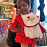 baby, child, smiling, red_clothing, bow, headband, bib, indoor, cafe, colorful, tablecloth, floral_pattern, person, happy, curly_hair, window, chair, hand, holding, toddler
