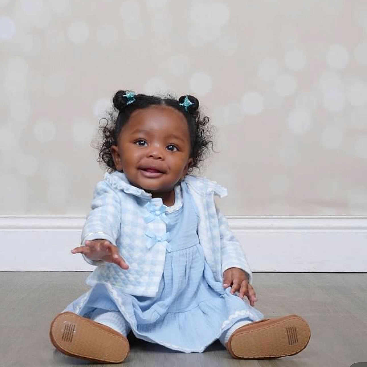 Sarai joined the competition — help win amazing prizes! toddler, child, smiling, sitting, blue_dress, hair_buns, indoor, floor, cute, baby, portrait, happy, reaching_hand, long_sleeves, wall, soft_background, shoes, young_child, person, clothing
