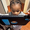 child, tablet, braided_hair, indoors, flooring, wall, technology, person, game, focus, young, standing, gray_clothing, feet, candid, device, hand, brown_floor, home, curiosity