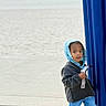child, boy, hoodie, blue_hoodie, jeans, sneakers, concrete, pole, blue_pole, beach, ocean, outdoor, sidewalk, casual_clothing, standing, curious, holding_object, daylight, urban, portrait