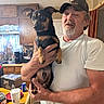 man, dog, person, portrait, indoors, kitchen, pet, pet_owner, holding, animal, black_and_tan, short_hair, cap, beard, white_shirt, wooden_cabinets, countertop, cup, soda_can, laundry_basket