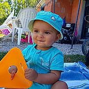 Rafaël a rejoint le concours — aidez-le/la à gagner de superbes lots ! baby, toddler, child, hat, blue_clothing, outdoor, plastic_chair, toy, orange_toy, blanket, grass, building, sunlight, summer, playful, smiling, sitting, person, daytime, casual