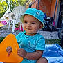 baby, toddler, child, hat, blue_clothing, outdoor, plastic_chair, toy, orange_toy, blanket, grass, building, sunlight, summer, playful, smiling, sitting, person, daytime, casual