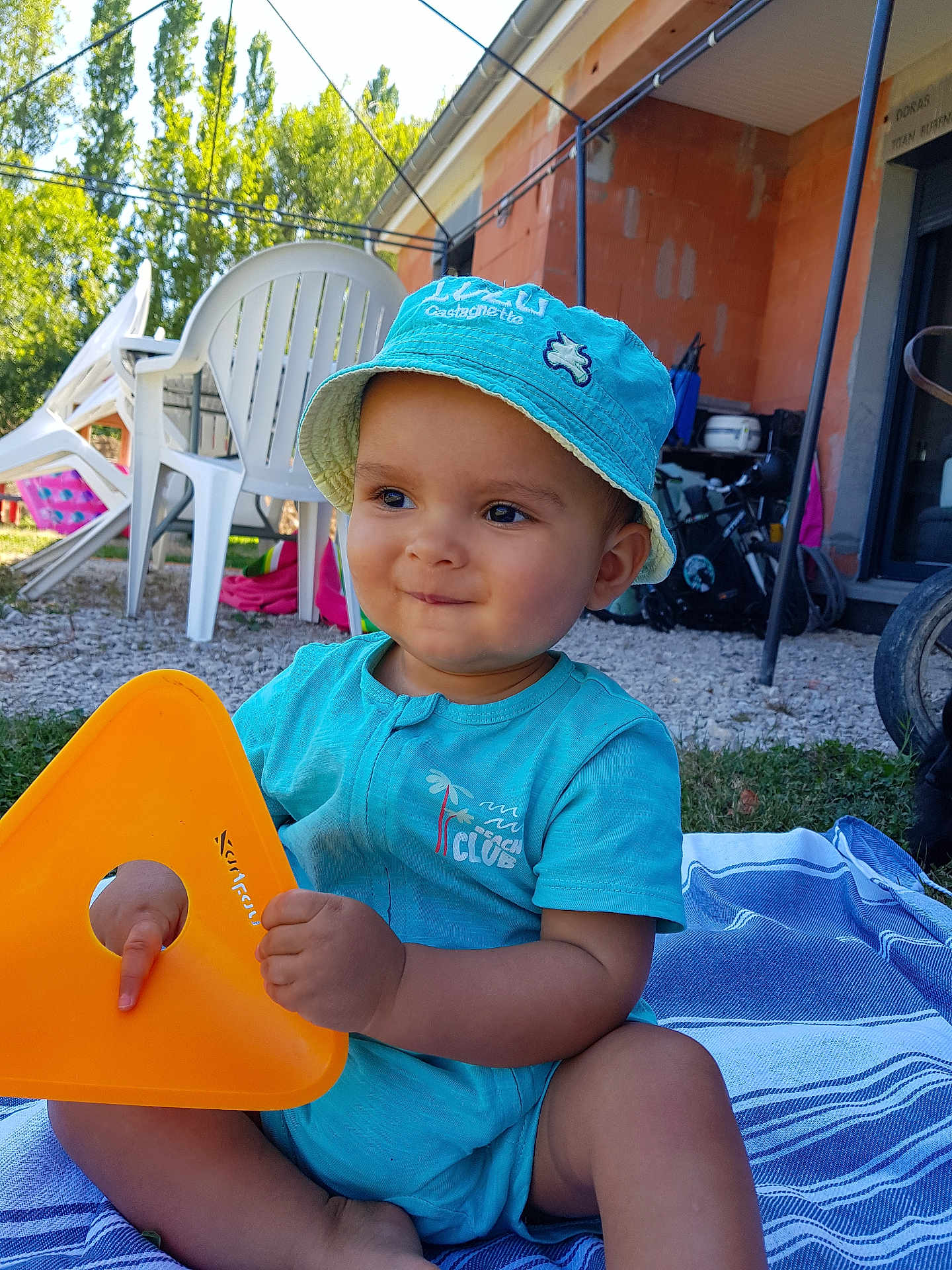 Rafaël a rejoint le concours — aidez-le/la à gagner de superbes lots ! baby, toddler, child, hat, blue_clothing, outdoor, plastic_chair, toy, orange_toy, blanket, grass, building, sunlight, summer, playful, smiling, sitting, person, daytime, casual