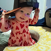 Lexi is registered to the contest to win money with this photo: toddler, child, baby, cap, hat, pink_dress, red_hearts, baby_seat, cushion, indoor, smiling, face, person, playful, cute, clothing, headwear, pattern, furniture, room