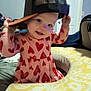 toddler, child, baby, cap, hat, pink_dress, red_hearts, baby_seat, cushion, indoor, smiling, face, person, playful, cute, clothing, headwear, pattern, furniture, room