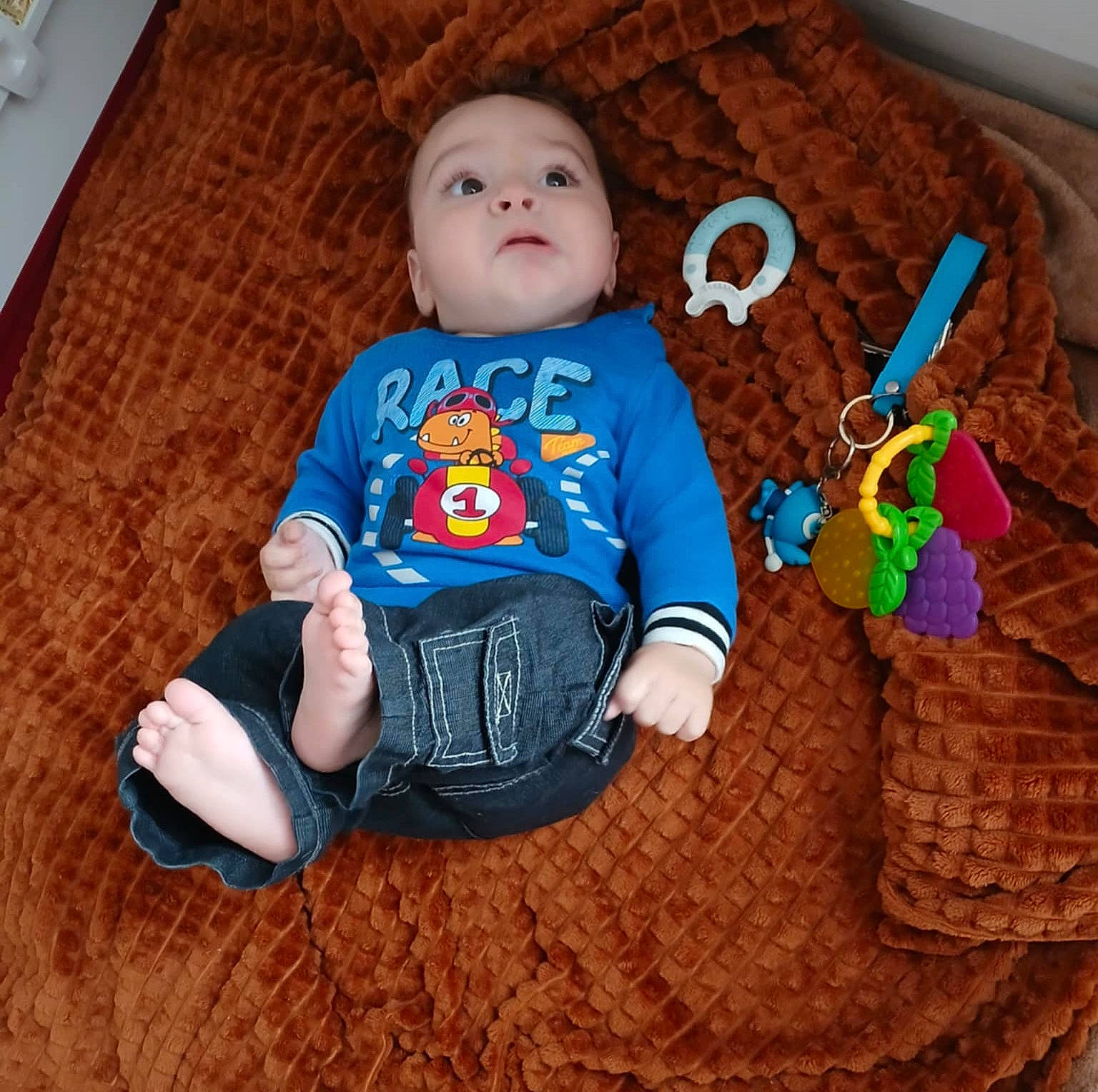 Nathan participe au concours pour gagner de l'argent avec cette photo : baby, barefoot, blanket, blue_shirt, brown_blanket, casual_clothing, child, colorful_toys, curious_expression, cute, denim_pants, indoors, infant, lying_down, person, relaxed, small_feet, soft_texture, teething_ring, toy