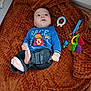 Nathan participe au concours pour gagner de l'argent avec cette photo : baby, infant, blue_shirt, denim_pants, barefoot, toy, teething_ring, blanket, brown_blanket, lying_down, curious_expression, indoors, child, person, cute, soft_texture, colorful_toys, casual_clothing, relaxed, small_feet