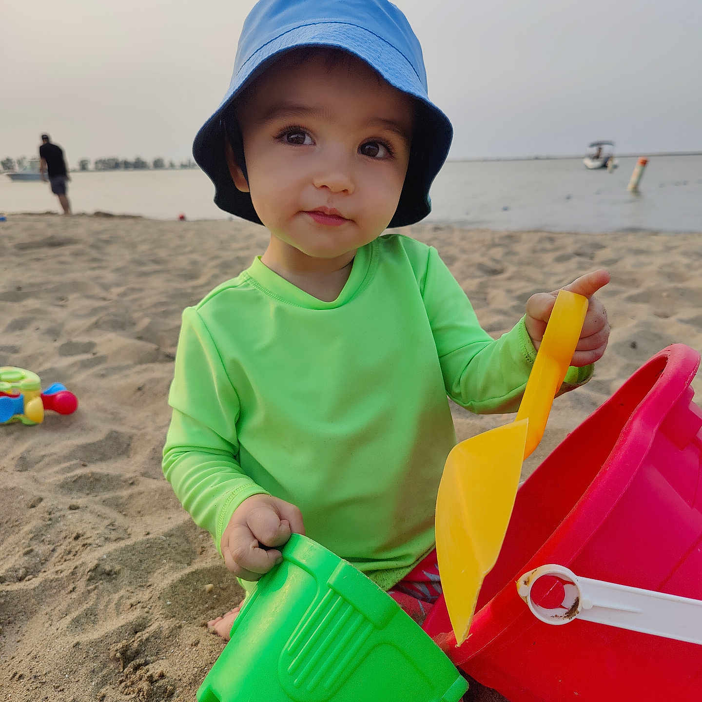 Luca joined the competition — help win amazing prizes! toddler, child, beach, sand, bucket, shovel, hat, green_clothing, toy, sea, water, boat, outdoor, play, summer, sunlight, person, recreation, vacation, sand_toys