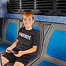 child, boy, smiling, seated, public_transport, bus, vehicle_interior, blue_seats, tablet, electronics, black_shirt, shorts, red_hair, portrait, indoors, travel, commuter, ventilation, casual_clothing, happy