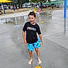 Maxwell joined the competition — help win amazing prizes! child, boy, girl, splash_pad, water, fountain, outdoor, summer, barefoot, wet, playground, park, picnic_table, umbrella, trees, street, buildings, black_shirt, blue_shorts, portrait