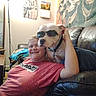 Mekkio is registered to the contest to win money with this photo: person, dog, sunglasses, couch, leather, indoor, tapestry, skull_art, wall_art, lamp, calendar, blanket, relaxed, smiling, casual_clothing, home, furniture, portrait, pet, companion