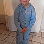child, boy, denim, jumpsuit, indoors, tile_floor, shoes, standing, casual_clothing, portrait, smiling, hands_clasped, young, light_skin, short_hair, buttoned_clothing, full_body, neutral_expression, home_interior, door