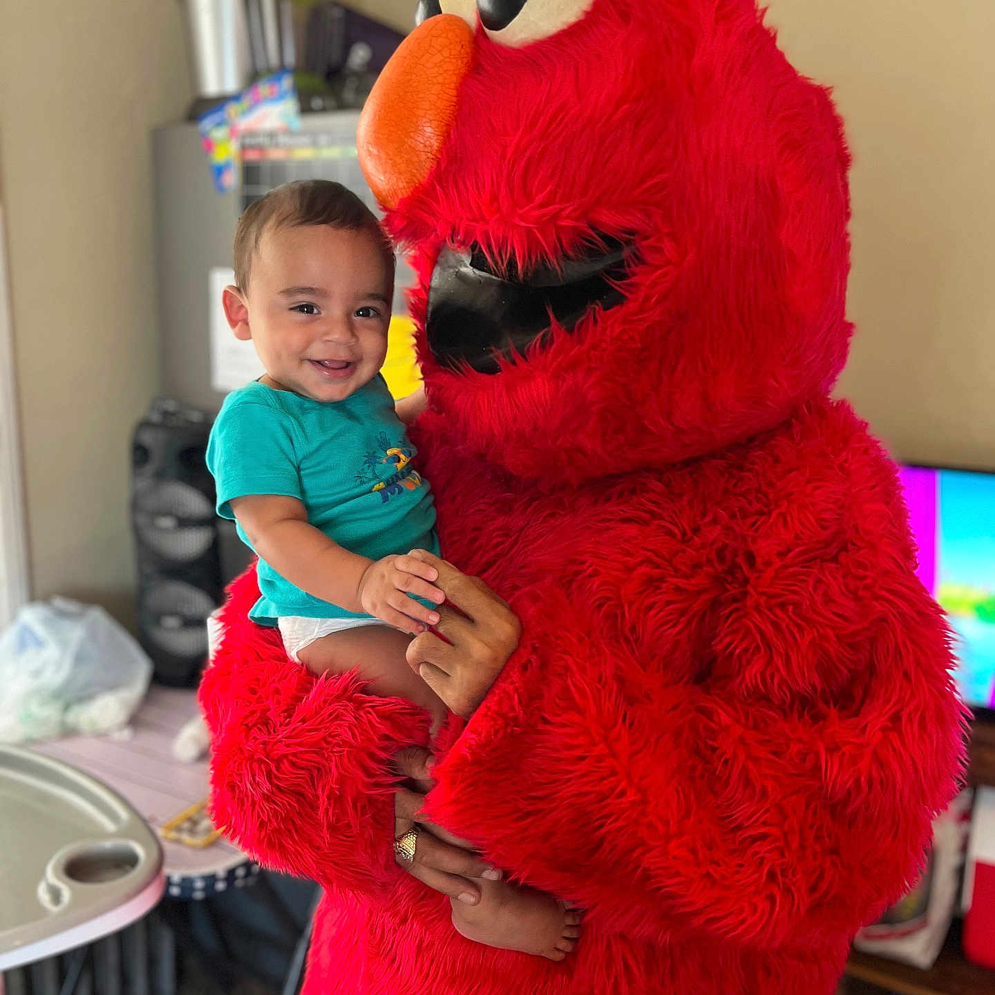 Messiah is registered to the contest to win money with this photo: baby, blue_shirt, child, costume, cute, elmo, face, foot, furniture, hand, happy, holding, indoor, living_room, person, red_fur, smiling, television, toddler, toy_character
