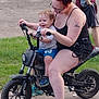 Edward is registered to the contest to win money with this photo: toddler, woman, motorbike, sidewalk, grass, boy, outdoor, casual_clothing, smiling, red_hair, shorts, sneakers, tattoo, glasses, summer, play, fun, child, adult, vehicle