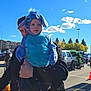 toddler, child, costume, blue, outdoor, sunny, sky, clouds, balloons, parking_lot, adult, person, holding, festival, event, trees, vehicle, black_jacket, cute, playful