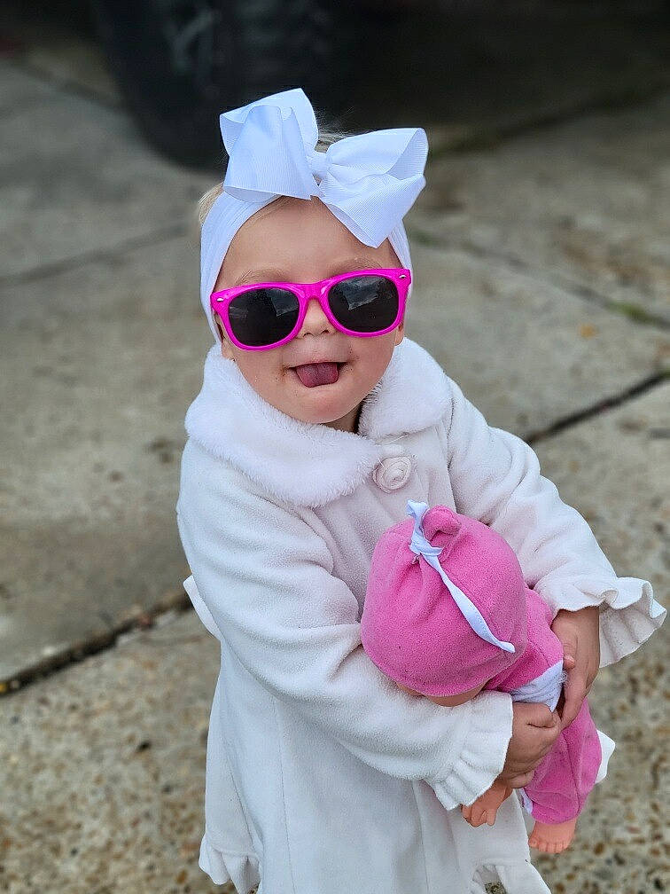 Amelia is registered to the contest to win money with this photo: baby_toddler_clothing, cap, eyewear, face, glasses, goggles, happy, head, headgear, leisure, magenta, person, personal_protective_equipment, pink, plant, recreation, sleeve, sunglasses, toddler, tree
