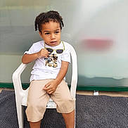 Fleury Nolan a rejoint le concours — aidez-le/la à gagner de superbes lots ! child, curly_hair, white_tshirt, camouflage, sunglasses, beige_shorts, white_shoes, plastic_chair, gray_carpet, indoor, sitting, looking_away, curious_expression, young_child, casual_clothing, shorts, footwear, person, furniture, background_blur