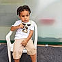 child, curly_hair, white_tshirt, camouflage, sunglasses, beige_shorts, white_shoes, plastic_chair, gray_carpet, indoor, sitting, looking_away, curious_expression, young_child, casual_clothing, shorts, footwear, person, furniture, background_blur