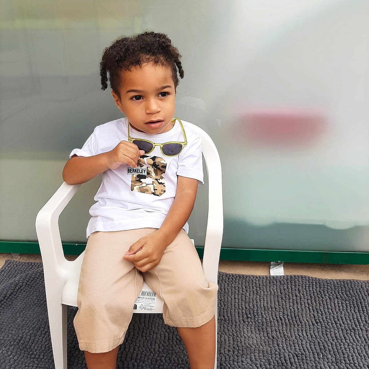 Fleury Nolan a rejoint le concours — aidez-le/la à gagner de superbes lots ! background_blur, beige_shorts, camouflage, casual_clothing, child, curious_expression, curly_hair, footwear, furniture, gray_carpet, indoor, looking_away, person, plastic_chair, shorts, sitting, sunglasses, white_shoes, white_tshirt, young_child