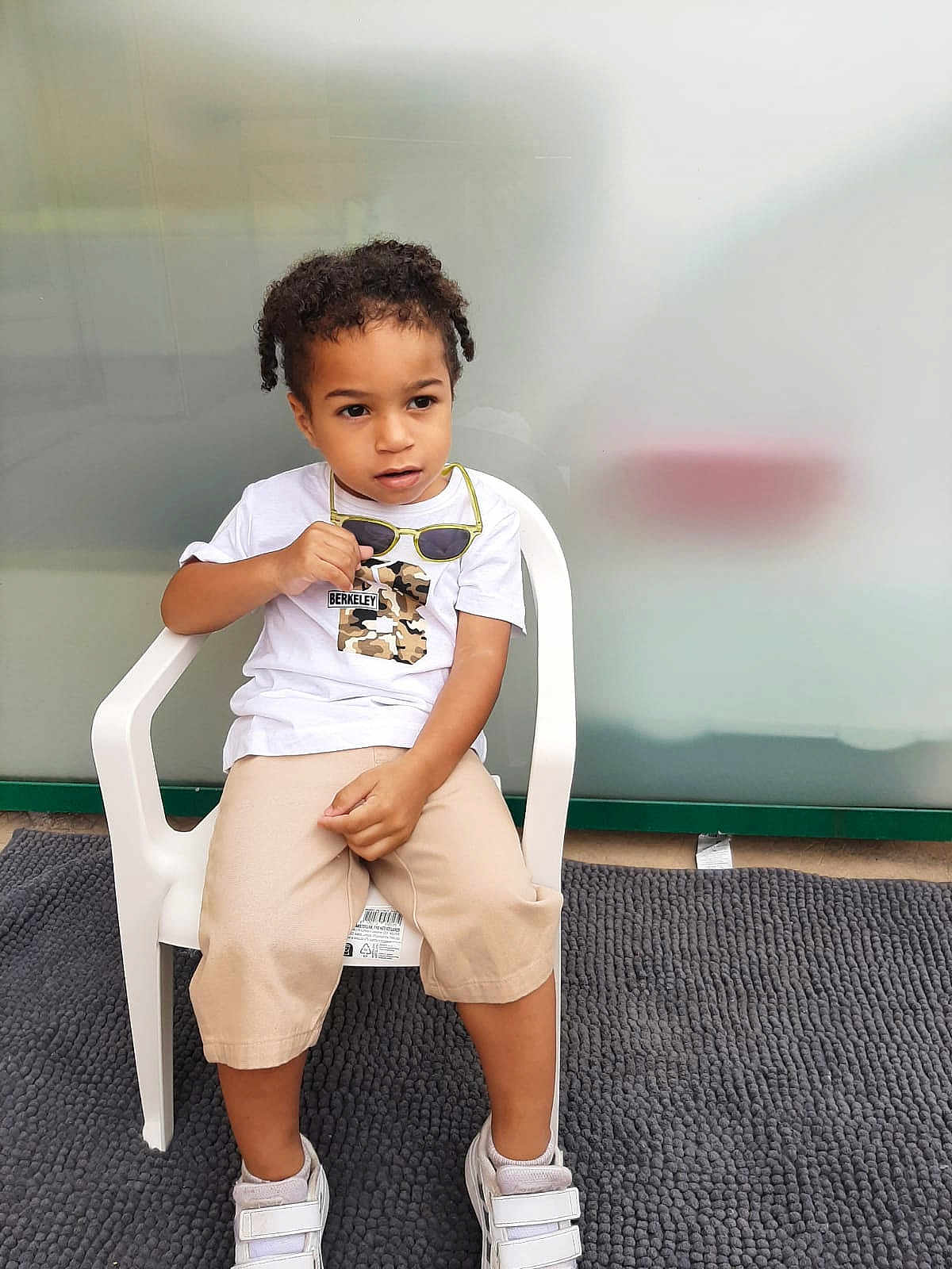 Fleury Nolan a rejoint le concours — aidez-le/la à gagner de superbes lots ! child, curly_hair, white_tshirt, camouflage, sunglasses, beige_shorts, white_shoes, plastic_chair, gray_carpet, indoor, sitting, looking_away, curious_expression, young_child, casual_clothing, shorts, footwear, person, furniture, background_blur
