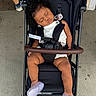 toddler, child, sleeping, stroller, curly_hair, white_outfit, floral_pattern, purple_shoes, footwear, seatbelt, outdoor, concrete, person, baby, nap, relaxed, black_stroller, casual, daytime, rest