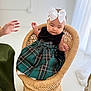 baby, child, wicker_chair, white_bow, headband, green_dress, plaid, sitting, indoor, hand, adult_hand, floor, minimalist, soft_lighting, woven, cute, portrait, infant, clothing, person
