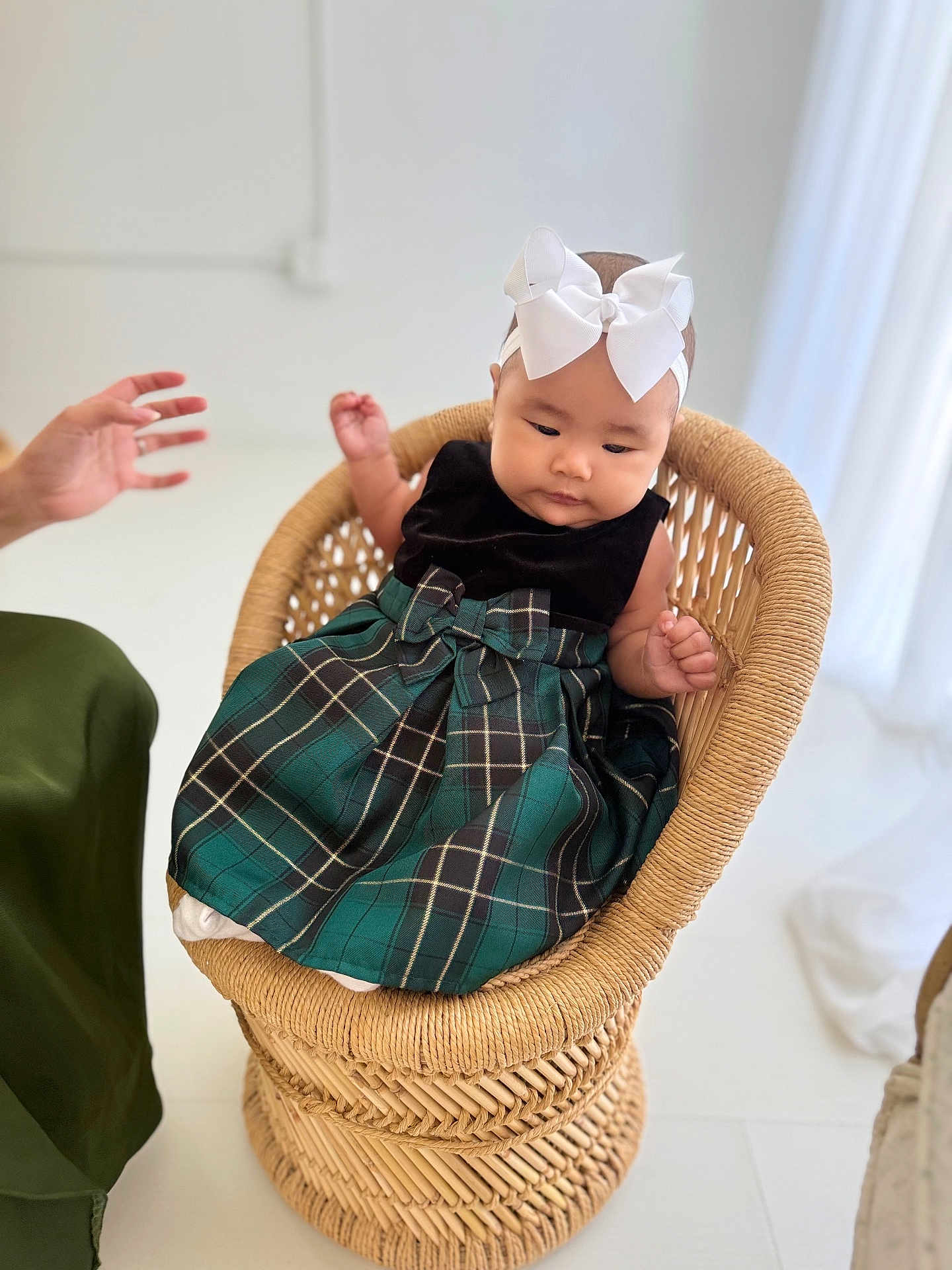 Ellie is registered to the contest to win money with this photo: baby, child, wicker_chair, white_bow, headband, green_dress, plaid, sitting, indoor, hand, adult_hand, floor, minimalist, soft_lighting, woven, cute, portrait, infant, clothing, person