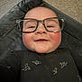 Aaron is registered to the contest to win money with this photo: baby, glasses, smile, face, child, black_clothing, origami_bird_pattern, lying_down, soft_texture, closeup, cheerful, indoors, infant, cute, head, eyewear, young_child, portrait, happy, wearing