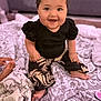 baby, smiling, child, sitting, blanket, patterned_clothing, indoor, happy, cute, infant, face, person, foot, hand, black_shirt, pants, cozy, soft_texture, home, cheerful