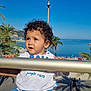 barefoot, beach, blue_sky, child, curly_hair, happy, holiday, nature, outdoor, palm_trees, person, portrait, railing, sea, shirt, shorts, summer, sunny, toddler, vacation
