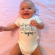 Valentino is registered to the contest to win money with this photo: baby, infant, smiling, onesie, text_on_clothing, pink_quilt, happy, cute, lying_down, child, portrait, skin, head, arms, legs, indoors, person, clothing, fabric, expression