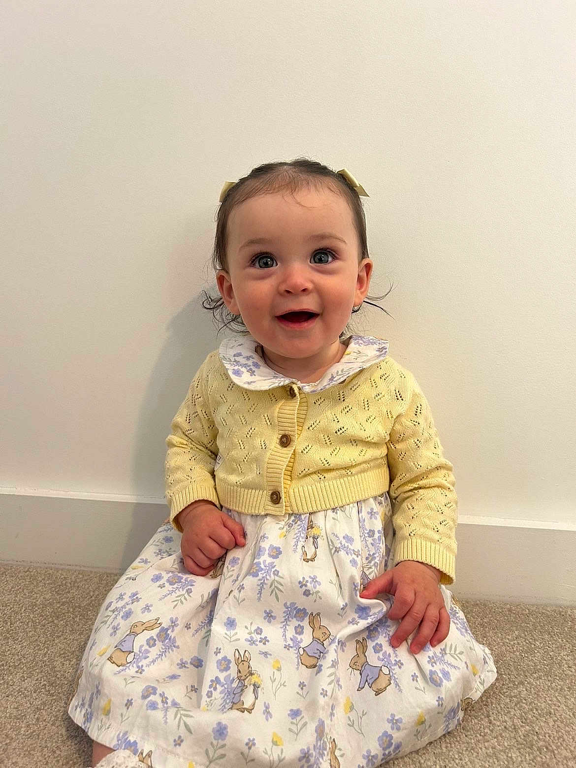Ayla joined the competition — help win amazing prizes! baby, child, girl, smiling, yellow_cardigan, floral_dress, bunny_prints, hair_bows, carpet, indoor, cute, happy, portrait, sitting, long_sleeves, wall, clothing, person, infant, toddler