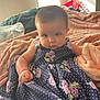 baby, child, dress, floral_pattern, blanket, indoor, person, portrait, cute, infant, hand, face, expression, sitting, cozy, soft, fabric, home, light, casual