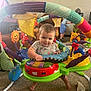 Leanna joined the competition — help win amazing prizes! baby, child, activity_jumper, toys, living_room, carpet, colorful, smiling, furniture, play, footwear, couch, person, indoor, happy, sock, blue_shorts, fan, plastic, seat