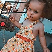 Alexis is registered to the contest to win money with this photo: toddler, child, curly_hair, dress, floral_pattern, outdoor, sidewalk, brick_wall, building, person, hand, footwear, sunlight, expression, stance, daytime, casual, young_child, portrait, holding
