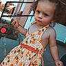 toddler, child, curly_hair, dress, floral_pattern, outdoor, sidewalk, brick_wall, building, person, hand, footwear, sunlight, expression, stance, daytime, casual, young_child, portrait, holding