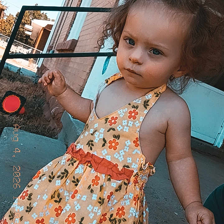 Alexis is registered to the contest to win money with this photo: toddler, child, curly_hair, dress, floral_pattern, outdoor, sidewalk, brick_wall, building, person, hand, footwear, sunlight, expression, stance, daytime, casual, young_child, portrait, holding