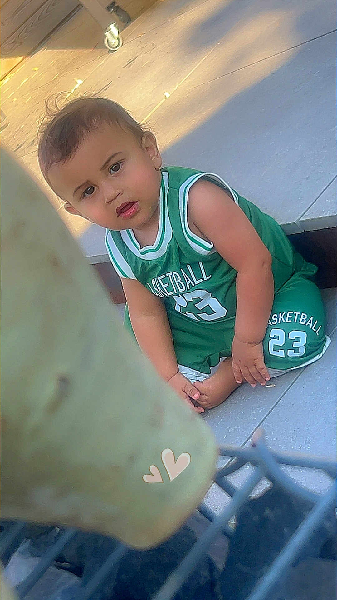 Alek participe au concours pour gagner de l'argent avec cette photo : child, toddler, basketball_jersey, green_clothing, sitting, floor, tiles, curious, young_child, outdoor, sunlight, heart_stickers, foreground_blur, person, casual, short_hair, barefoot, expression, daytime, sportswear