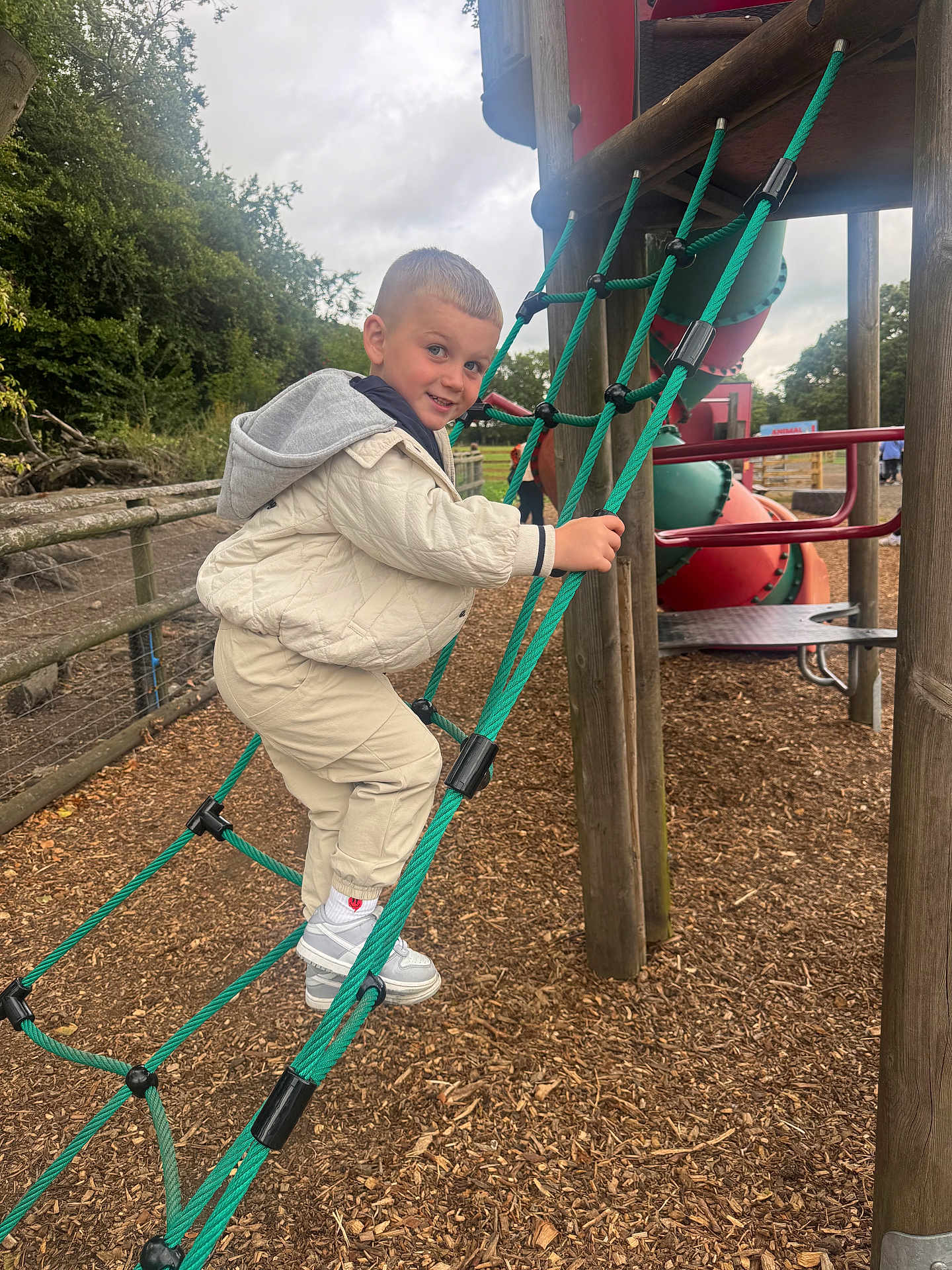 Jace is registered to the contest to win money with this photo: child, boy, playground, rope_ladder, climbing, outdoor, park, woodchips, jacket, pants, sneakers, smiling, fence, trees, play_equipment, daytime, happy, person, activity, casual_clothes