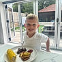 child, boy, freckles, white_polo, smiling, plate, corn_on_the_cob, steak, potato_wedges, ketchup, food, meal, kitchen, marble_countertop, glass_doors, backyard, outdoor_view, table, indoor, window