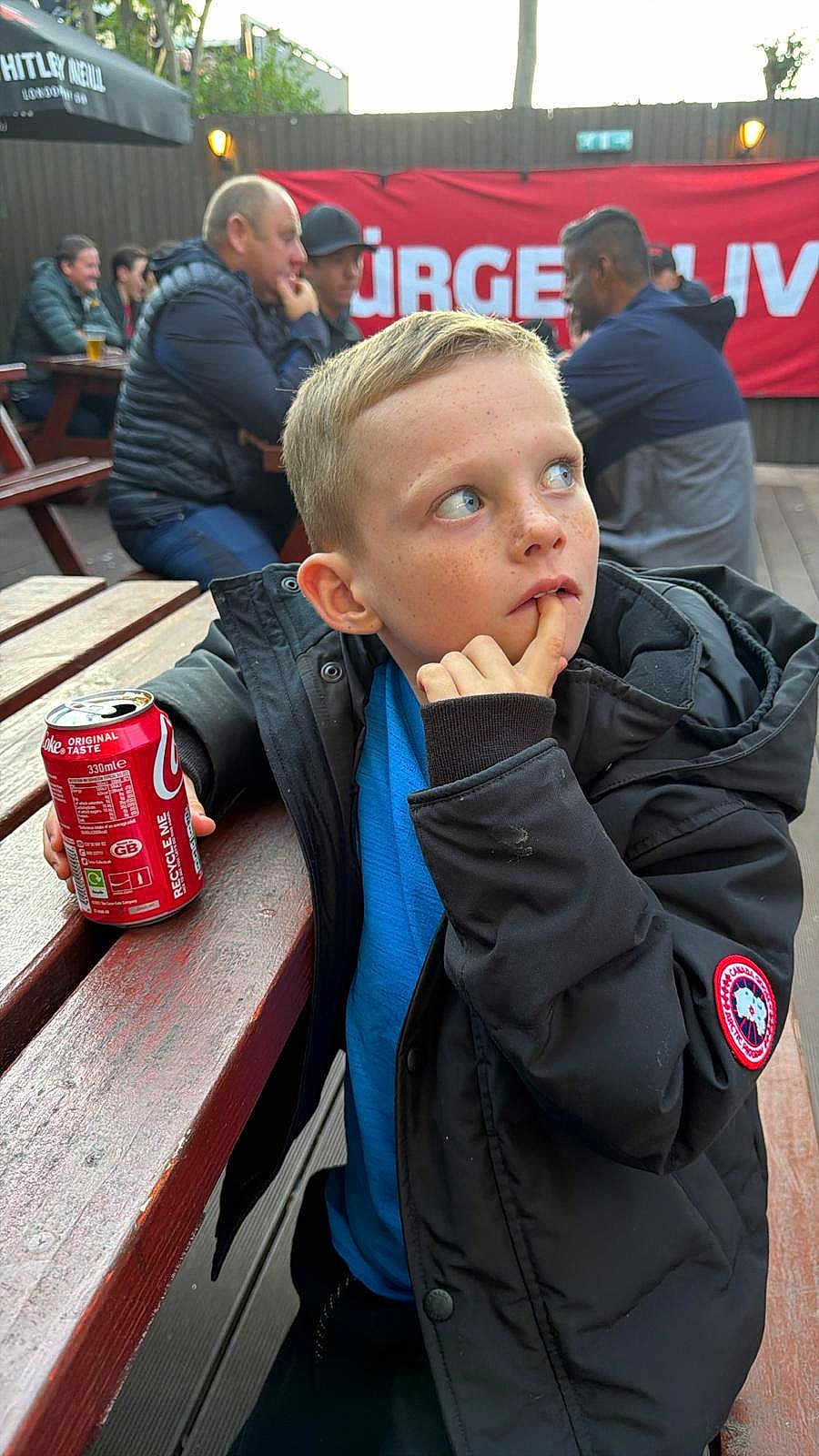 Jaxon is registered to the contest to win money with this photo: child, boy, freckles, outdoor, picnic_table, coke_can, black_jacket, blue_shirt, people, social_gathering, bench, casual, daylight, thoughtful, hand_near_mouth, beverage, wooden_table, jacket, freckled_face, curious