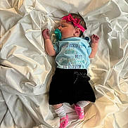 Janet is registered to the contest to win money with this photo: newborn, baby, sleeping, pacifier, headband, pink_bow, blue_shirt, black_shorts, white_sheet, sock, infant, clothing, lying_down, peaceful, cute, indoors, soft_texture, fabric, small_hands, small_feet