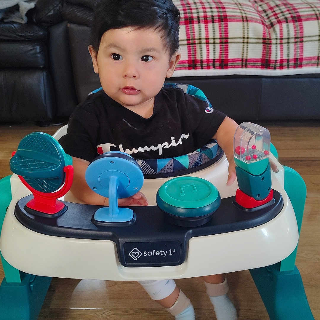 Eithan Gomez joined the competition — help win amazing prizes! baby, black_shirt, child, couch, curious, cute, floor, furniture, home, indoor, living_room, person, plaid_blanket, playtime, seated, socks, toddler, toy, walker, wood_floor