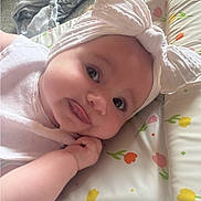 Riley is registered to the contest to win money with this photo: baby, infant, headband, tongue_out, cute, closeup, face, hand, floral_pattern, cushion, indoors, carpet, clothing, skin, lying_down, expression, child, playful, home, soft_lighting