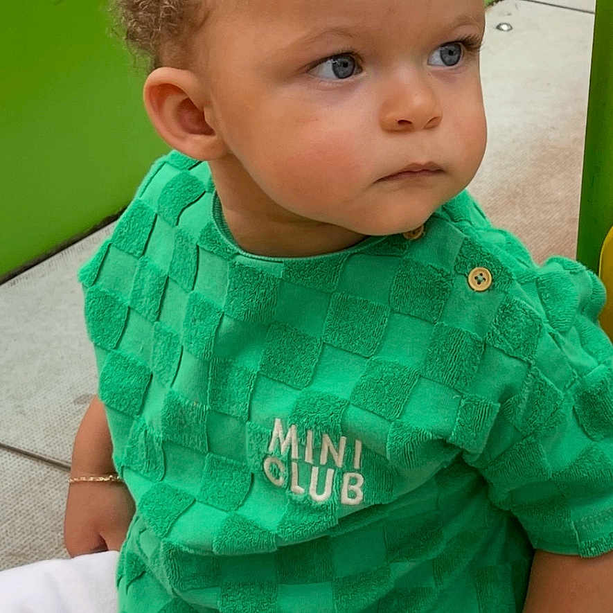 Siya a rejoint le concours — aidez-le/la à gagner de superbes lots ! blue_eyes, bracelet, casual_clothing, checkered_pattern, child, curious, curly_hair, cute, green_background, green_shirt, mini_club, outdoor, person, play_area, playground, sitting, thoughtful, toddler, white_pants, young_child
