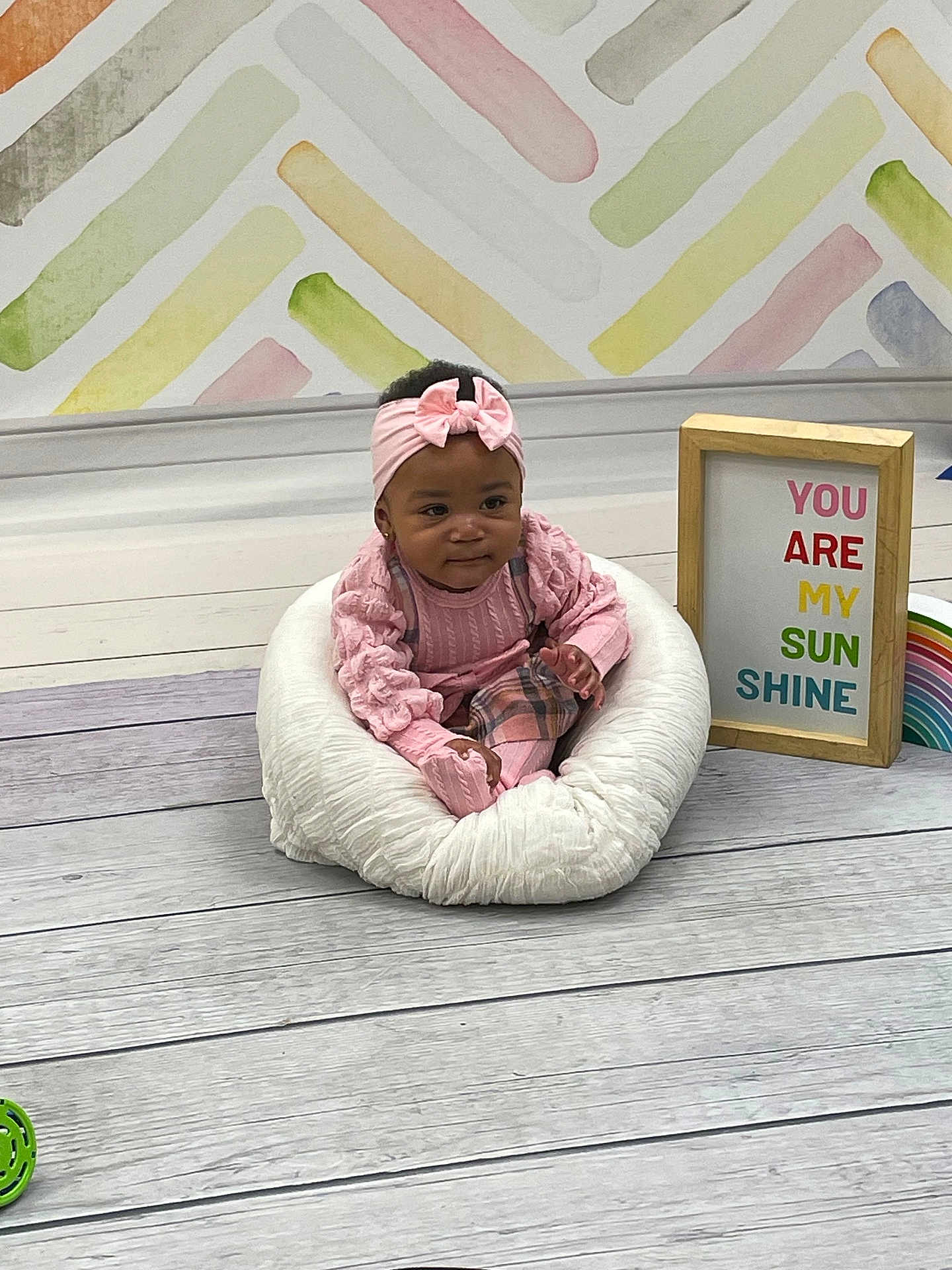 Miyori joined the competition — help win amazing prizes! baby, child, pink_clothing, headband, cushion, floor, wooden_floor, wall, colorful_wall, sign, text, rainbow, indoor, portrait, sitting, infant, smiling, decor, frame, cute