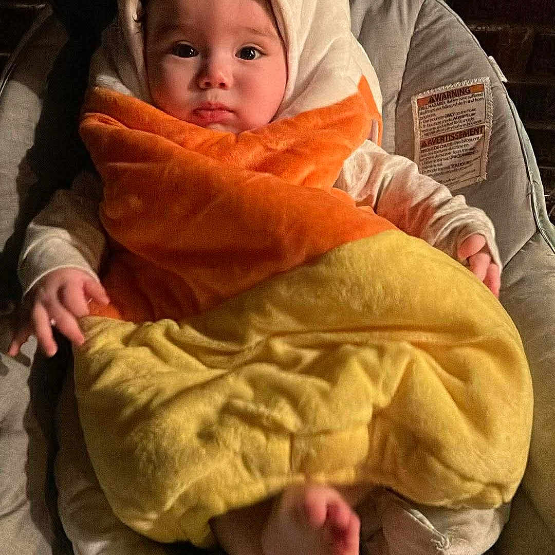 Nolan joined the competition — help win amazing prizes! baby, infant, child, portrait, swaddled, blanket, orange_blanket, yellow_blanket, hood, baby_seat, sitting, face, foot, cozy, indoor, brick_wall, warm_lighting, adorable, looking_at_camera, close_up